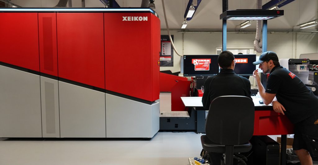 The diagonally red and grey coloured Xeikon printer, redesigned by Achilles Design, placed in a working environment with two employees.